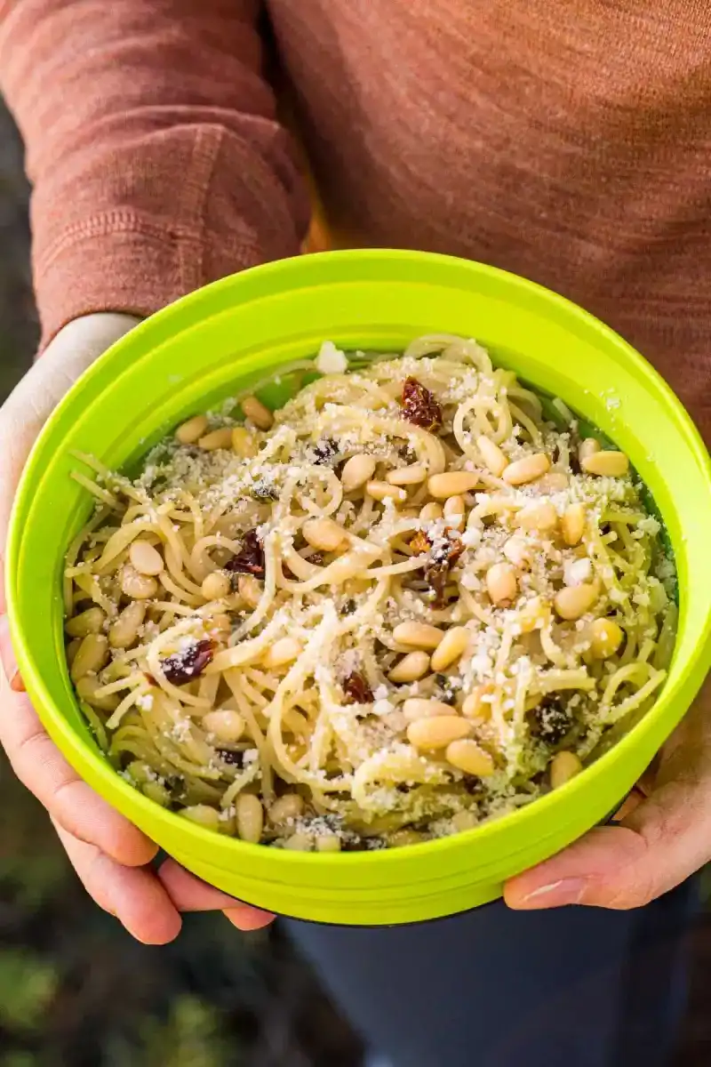   En person, der holder en skål fyldt med pasta, pestosauce, pinjekerner og soltørrede tomater.