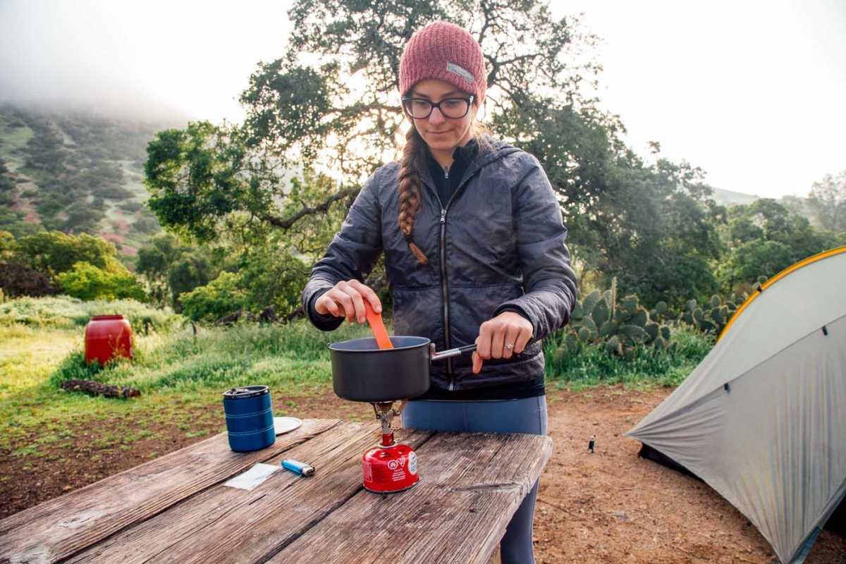 Kvinne som lager mat over en backpacking-komfyr med et telt i bakgrunnen.