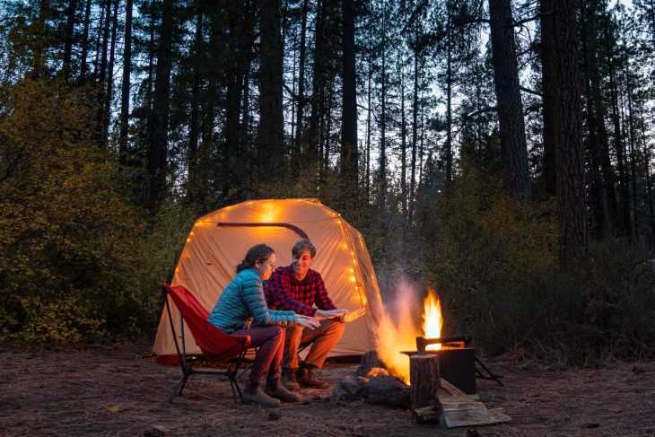 Меган и Майкъл седят близо до лагерен огън с къмпинг палатка на заден план