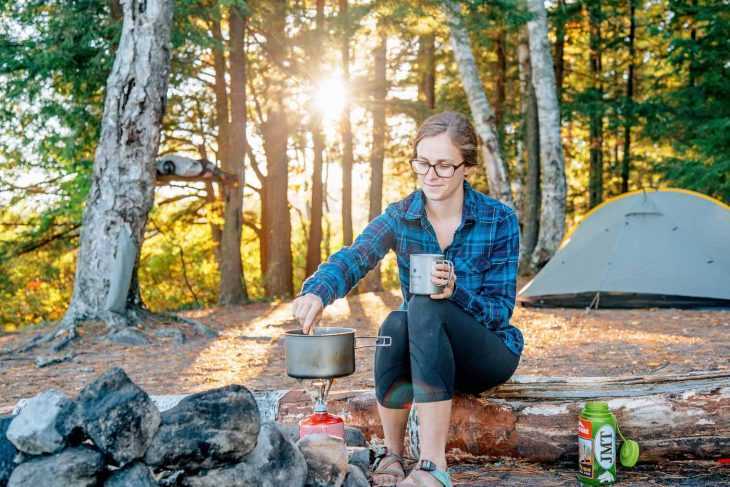 Megan sitzt auf einem Felsen neben einem Lagerfeuer und rührt in einem Topf mit Essen