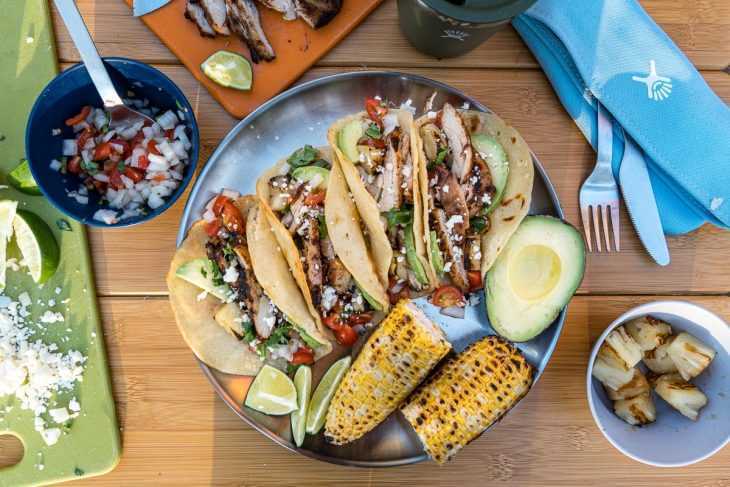 Quatre tacos disposés sur une assiette avec des épis de maïs grillés et un demi-avocat