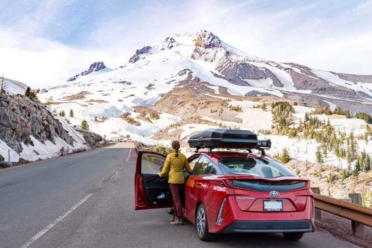 En röd bil står parkerad på platsen för en väg. Megan kliver ut ur bilen för att titta på Mt. Hood.