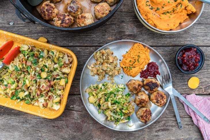 O farfurie umplută cu chifteluțe, varză de Bruxelles și cartofi dulci pe o masă înconjurată de feluri de mâncare