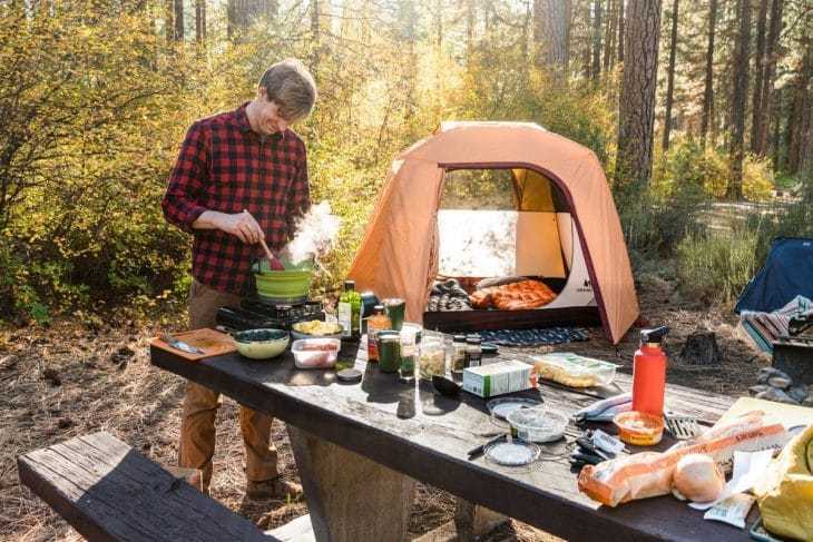 キャンプテーブルに立って鍋のトルテッリーニスープをかき混ぜるマイケル。キャンプの風景が背景にあります