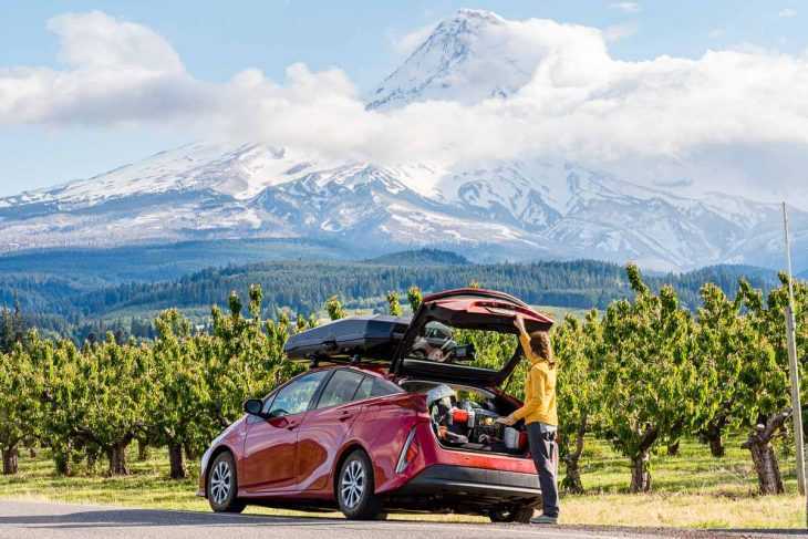 Меган открывает багажник красной машины, припаркованной на обочине дороги рядом с фруктовым садом. Вдалеке виднеется гора Худ, окутанная облаками.