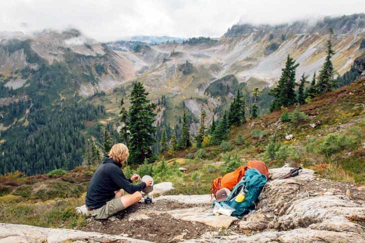 Mand laver et backpacking-måltid med bjerge i baggrunden.