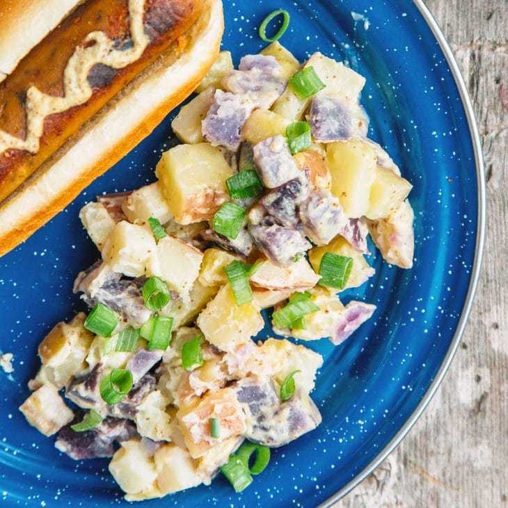 Různobarevný bramborový salát vedle párku v rohlíku na modrém talíři.