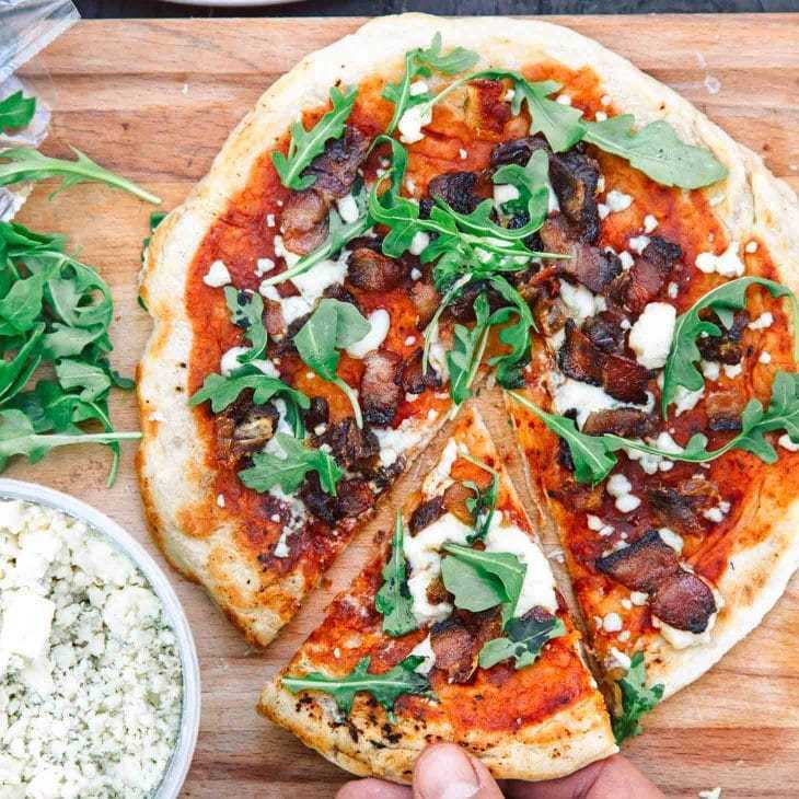 Sebuah tangan meraih sepotong pizza yang baru dibuat dengan saus tomat, keju, arugula, dan potongan daging asap di atas talenan kayu.