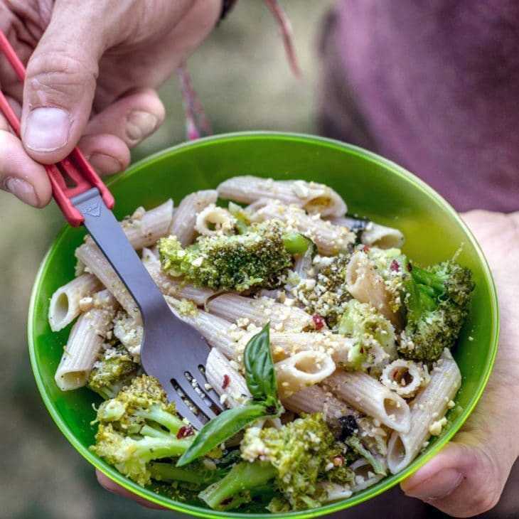 Eine Person isst im Freien mit einer Gabel einen gesunden Brokkoli-Nudel-Salat aus einer grünen Schüssel.