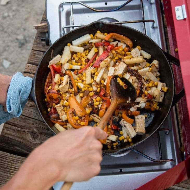 Mešanje mešanice pisane zelenjave in tofuja v ponvi nad taborniškim štedilnikom, ki ujame bistvo okusnega kuhanja na prostem.