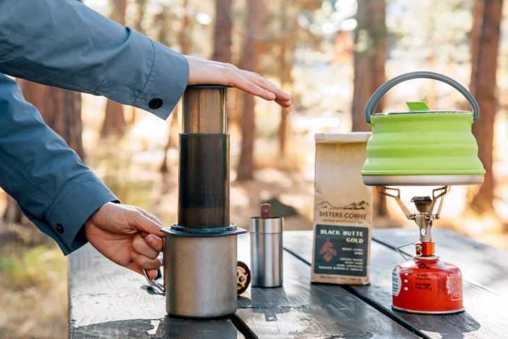 ميغان تستخدم آلة الإيروبريس لتحضير فنجان من القهوة. يوجد موقد مخيم وغلاية في الإطار على الطاولة.