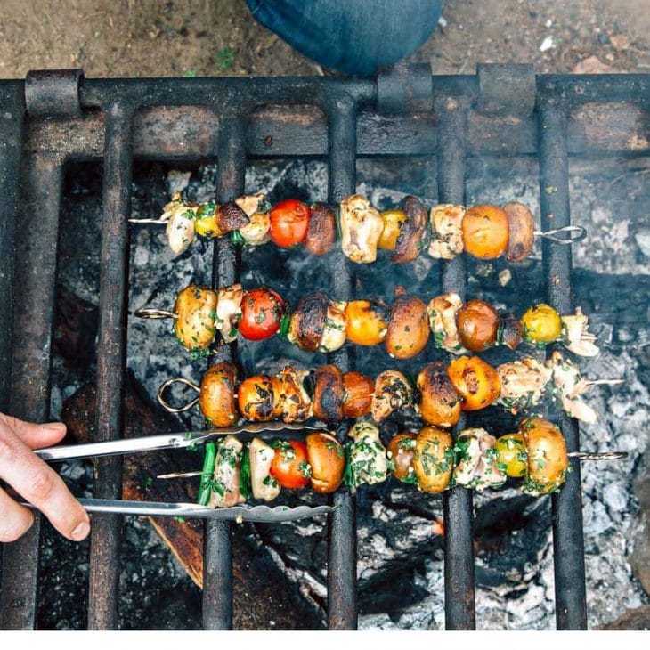 Bunte Spieße mit gemischtem Gemüse über offener Flamme grillen, wobei eine Person sie mit einer Zange dreht, um ein gleichmäßiges Garen zu gewährleisten.