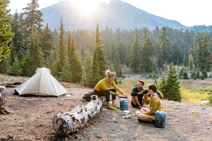 Tiga orang sedang memasak di samping tenda backpacking dengan gunung di kejauhan