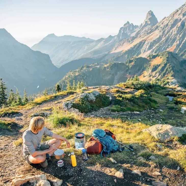 Michael sırt çantasıyla seyahat ettiği yemek pişirme seti ve arka planda Cascade dağlarıyla oturuyor