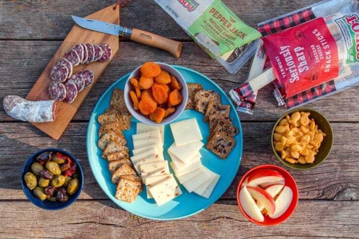 Ost, frugt, salami, nødder og kiks vist på et campingbord.