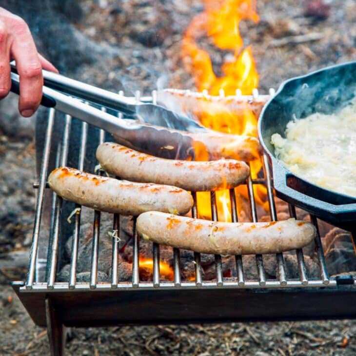 Grătirea cârnaților la foc deschis cu ceapa călită într-o tigaie din apropiere, captând esența gătitului în aer liber.