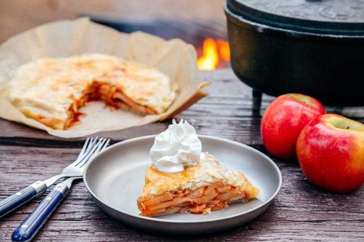 Hiwa ng apple pie sa isang plato na may Dutch oven at campfire sa background