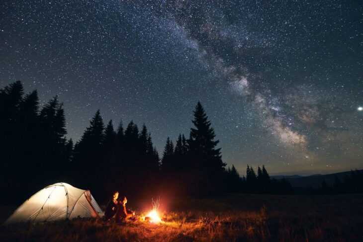 Um casal sentado ao lado de uma barraca e de uma fogueira sob um céu estrelado
