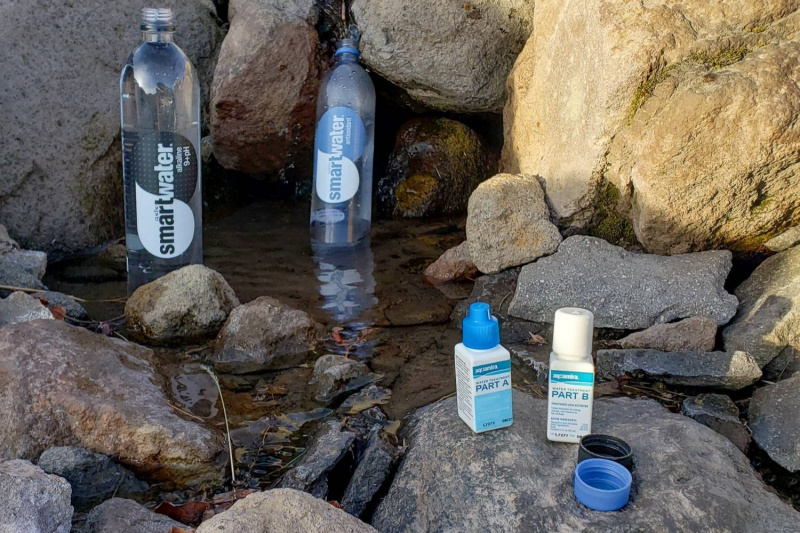   Aquamira pudeles sēž uz akmens avota priekšā ar divām ūdens pudelēm