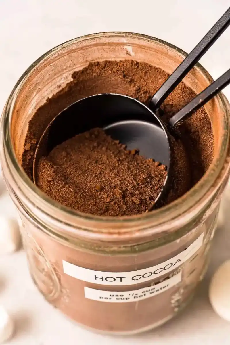   Een bovenaanzicht van een stenen pot vol warme chocolademelkmix met een maatbeker van ¼ kopje die de mix eruit haalt voor zelfgemaakte warme chocolademelk.