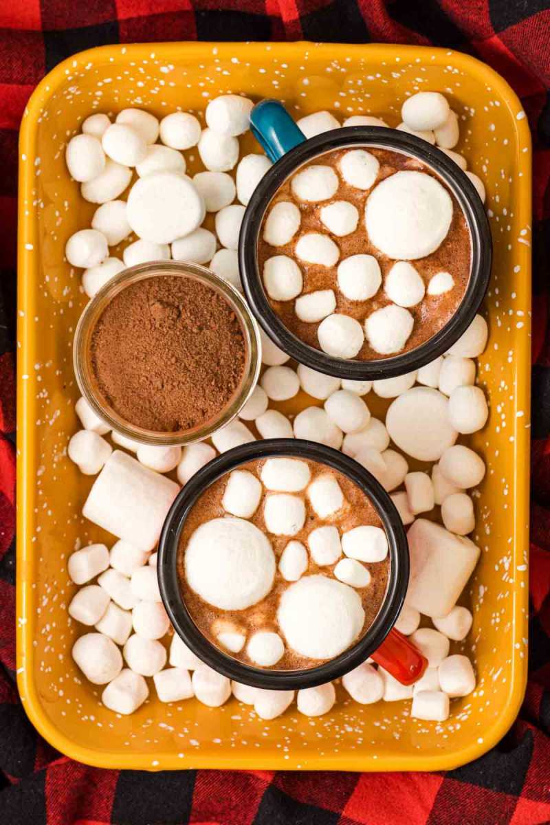   Două căni de cacao fierbinte cu vârf de marshmallow, una într-o cană albastră și una într-o cană roșie, pe o tavă galbenă cu un borcan de amestec fierbinte de cacao și mai multe bezele.