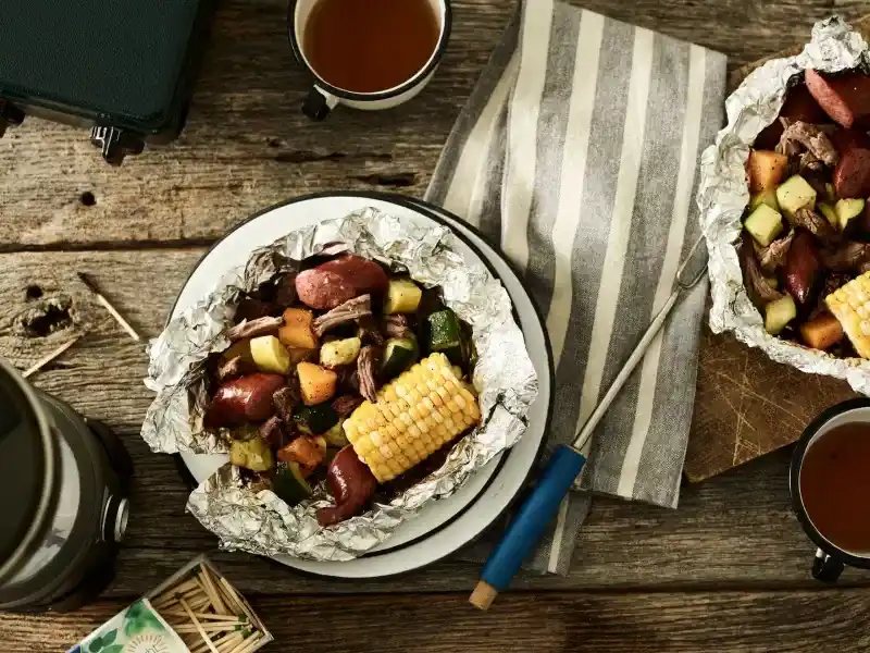   لکڑی کے کیمپ کی میز پر ورق کے پیکٹ میں سبزیوں کے ساتھ گائے کا گوشت۔