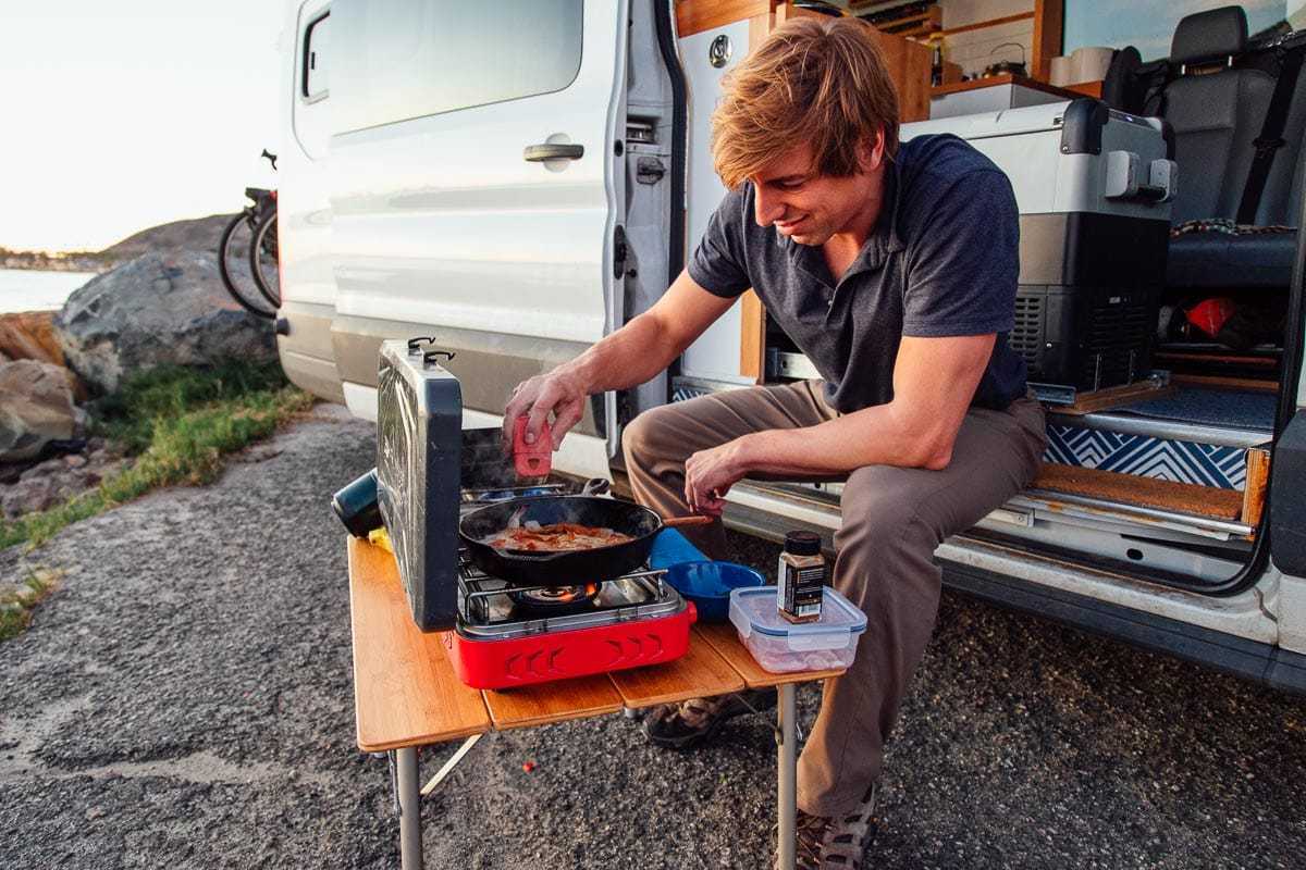 Michael está sentado na varanda de um trailer e cozinha em um fogão de acampamento.