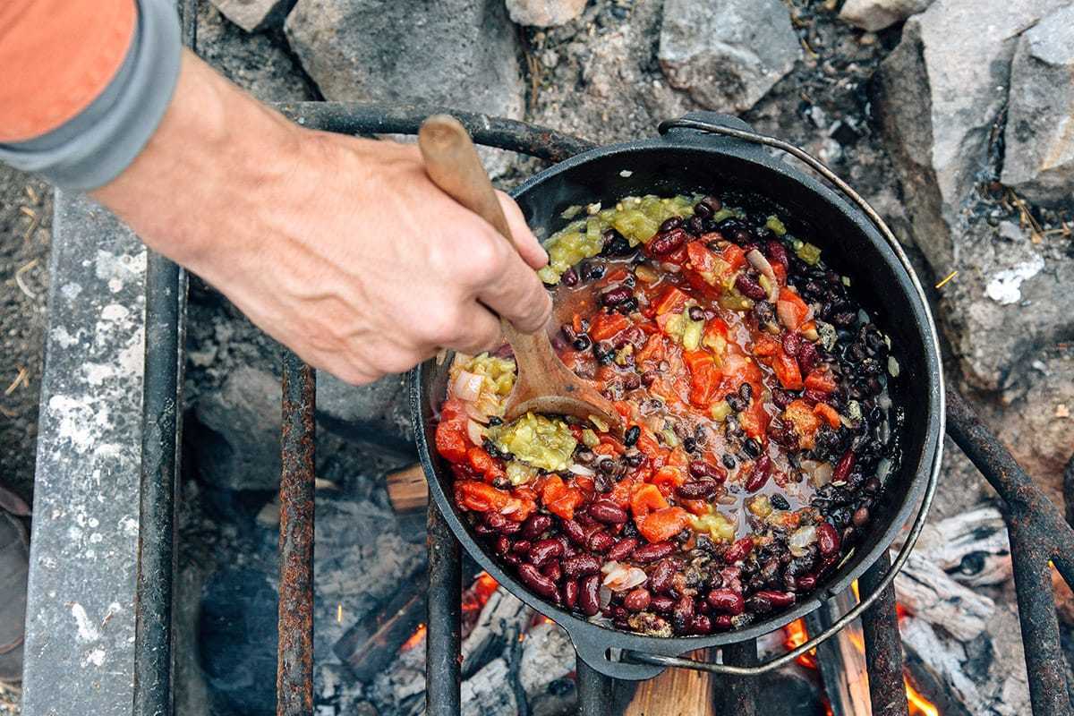 キャンプファイヤーの上のダッチオーブンでトマト、豆、唐辛子をかき混ぜるマイケル