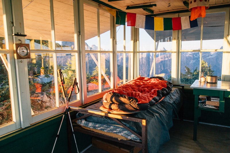   L'intérieur d'un belvédère d'incendie, avec un sac de couchage sur un lit et des fenêtres donnant sur une vue sur les sommets des montagnes.