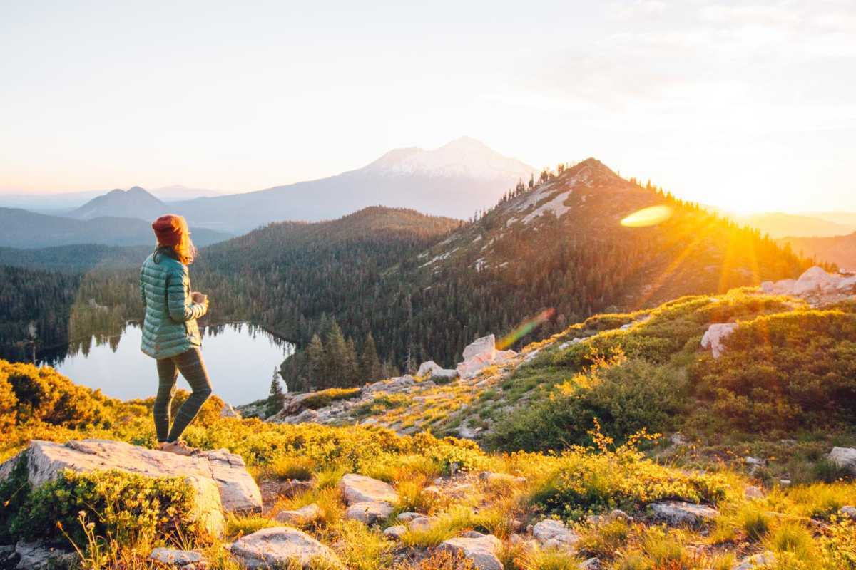 Megan está de pé segurando uma xícara de café, ela está olhando para o sol nascendo e o Monte Shasta ao longe