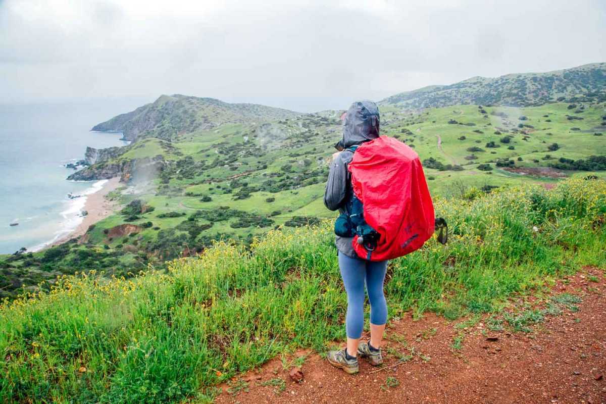 Kobieta na szlaku z widokiem na wyspę i ocean. Niebo jest deszczowe, a ona ma na sobie kurtkę przeciwdeszczową.