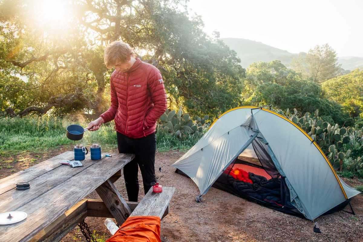 Človek pripravlja taborniško kavo za mizo za piknik s šotorom v ozadju