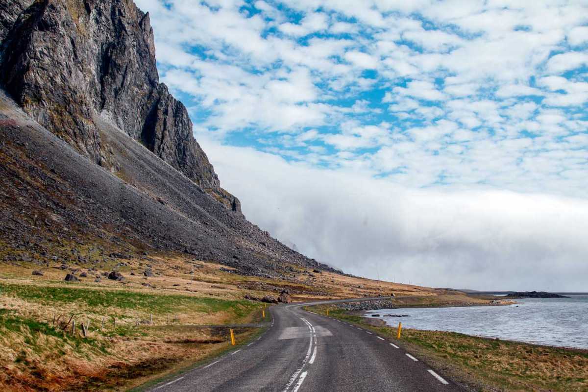 Een lege weg in IJsland. Er zijn zwarte bergen aan de linkerkant en de oceaan aan de rechterkant.