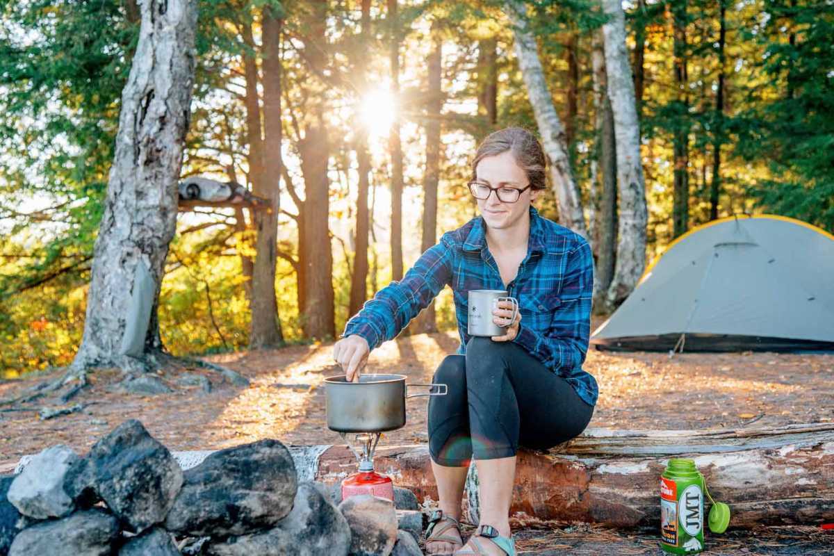Megan kamp ateşinin yanında bir kayanın üzerinde oturuyor ve bir tencere yemeği karıştırıyor