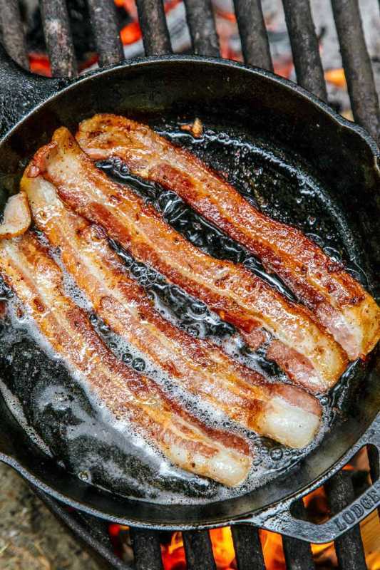 Quatro fios de bacon fritos em uma frigideira de ferro fundido sobre uma fogueira.