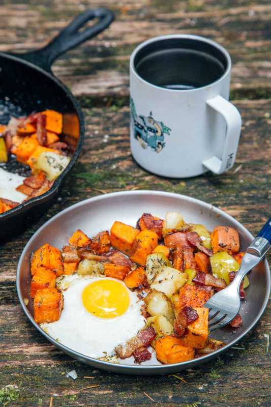 Hash apel dan ubi jalar & telur goreng di piring perak di meja perkemahan dengan secangkir kopi di latar belakang.