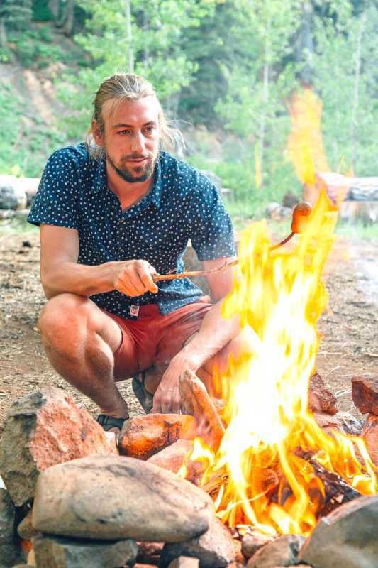 Nakayuko si Michael sa tabi ng campfire na nag-ihaw ng hotdog sa isang stick