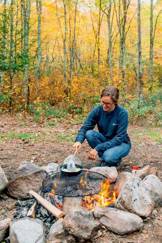 Megan cozinhando em uma fogueira com folhagem de outono ao fundo