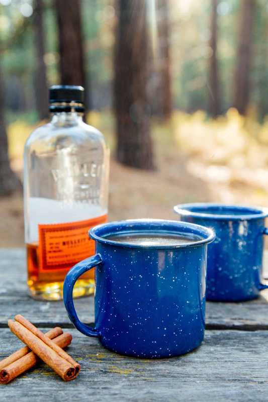 Scène forestière avec deux tasses de camping de cidre de pomme enrichi avec une bouteille de bourbon en arrière-plan.