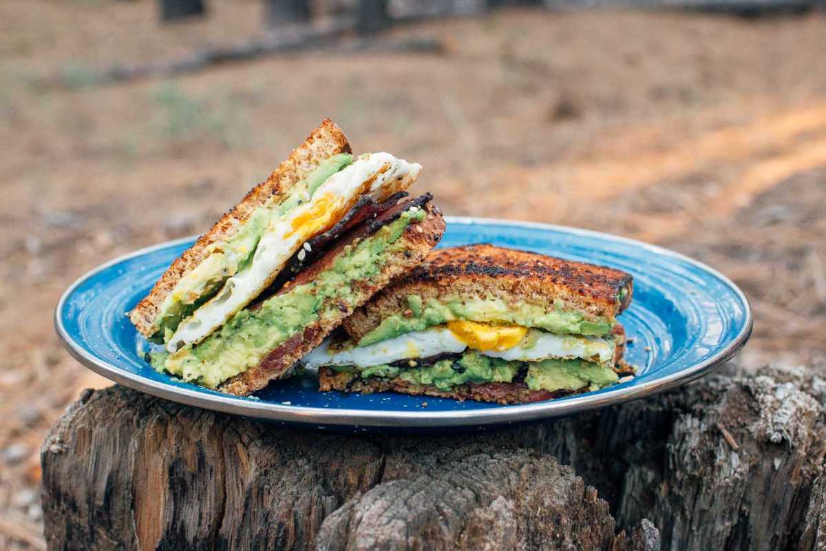 통나무 위에 놓인 파란색 캠핑 접시에 아보카도, 달걀, 베이컨을 곁들인 아침 샌드위치