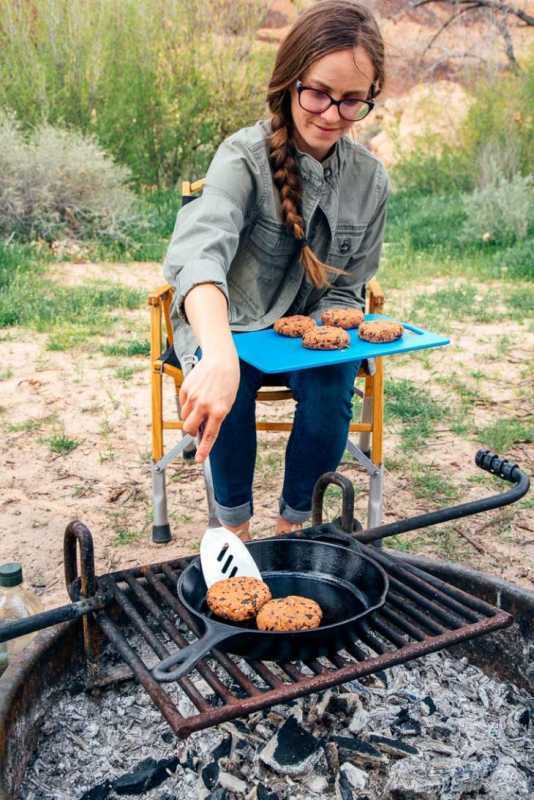 Megan brengt zwarte bonenburgerpasteitjes van een snijplank naar een koekenpan boven het kampvuur.