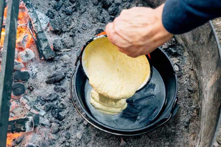 Michael despejando massa de pão de milho no forno holandês em uma fogueira