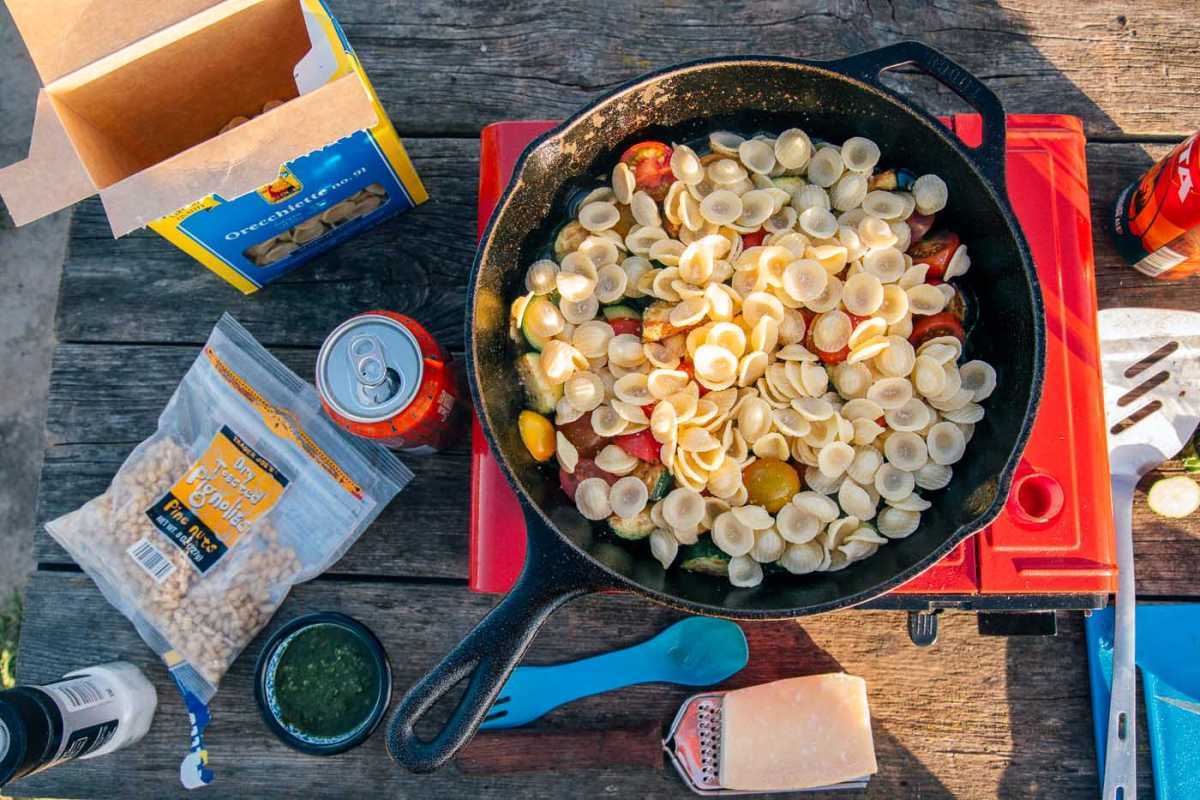 Oversikt over pasta i en støpejernsgryte på en campingkomfyr. Andre pastaingredienser som ost, pesto og pinjekjerner er på bordet.