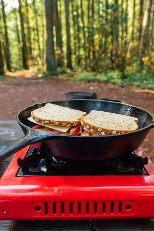 Ant grotelių kepti sūrio sumuštiniai keptuvėje ant stovyklavietės viryklės su mišku fone