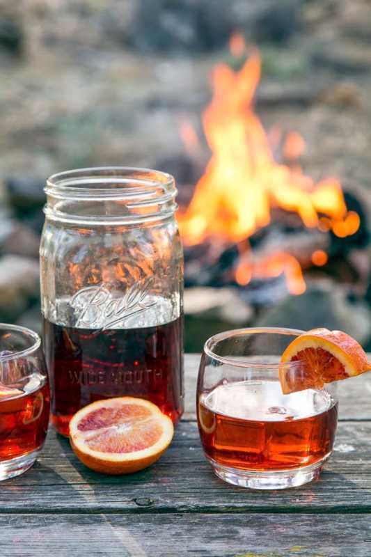 Cocktail Negroni într-un borcan de zidărie pe o masă în fața unui foc de tabără.