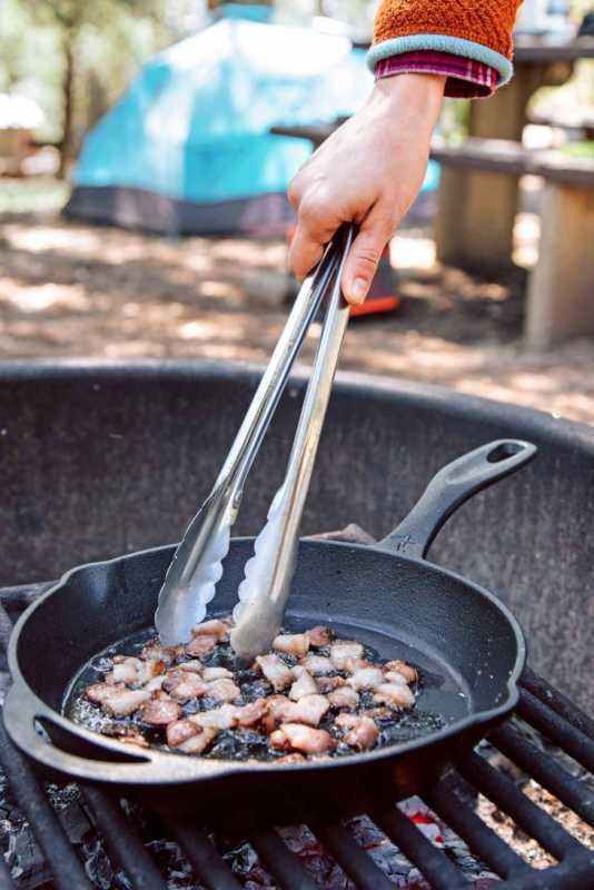 キャンプファイヤーの上でトングを使って、鋳鉄製のフライパンで刻んだベーコンを調理するミーガンさん