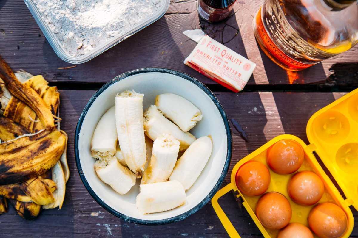 Ingredienser til bananbrød spredt på et campingbord: bananer i en bolle, egg, smør og mel.