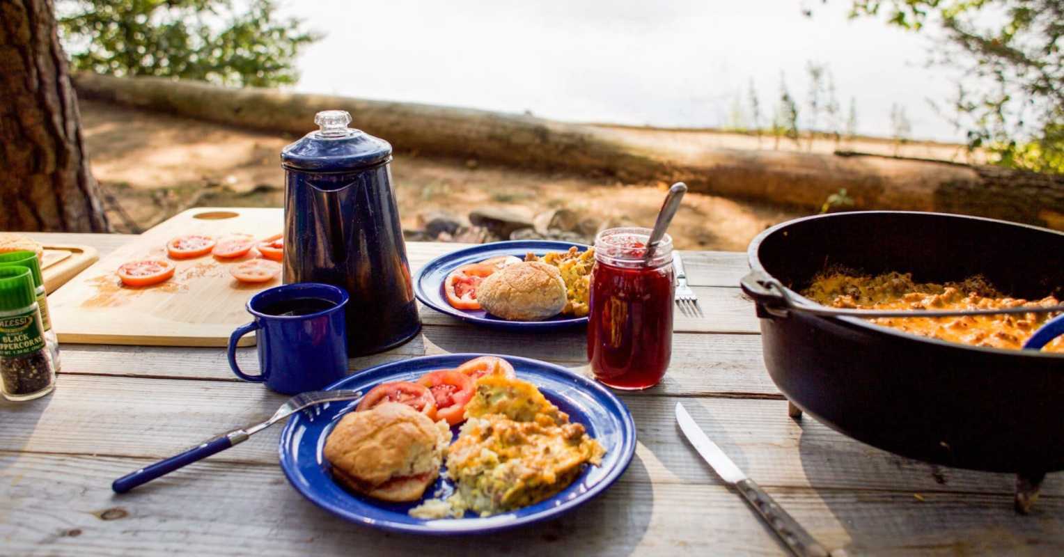कॉफ़ी के पॉट, अंडे के नाश्ते के साथ नीली प्लेट और फ़्रेम में डच ओवन के साथ एक शिविर दृश्य