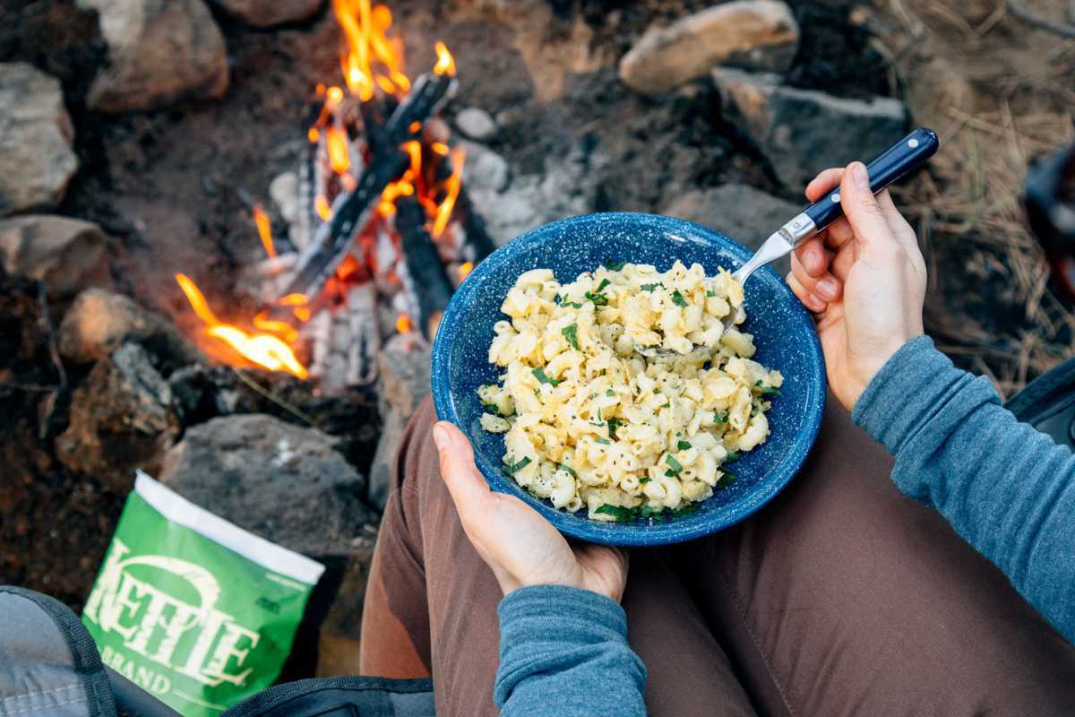 Mac en kaas in een blauwe kampeerkom naast een vuur.
