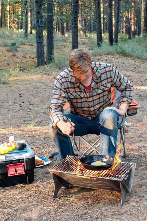 キャンプファイヤーの隣に座って鋳鉄のフライパンで卵を調理している男性